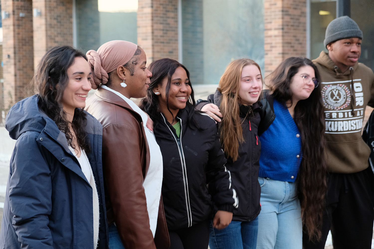 Six students stand shoulder to shoulder with arms around each other outside the campus on a cool day.
