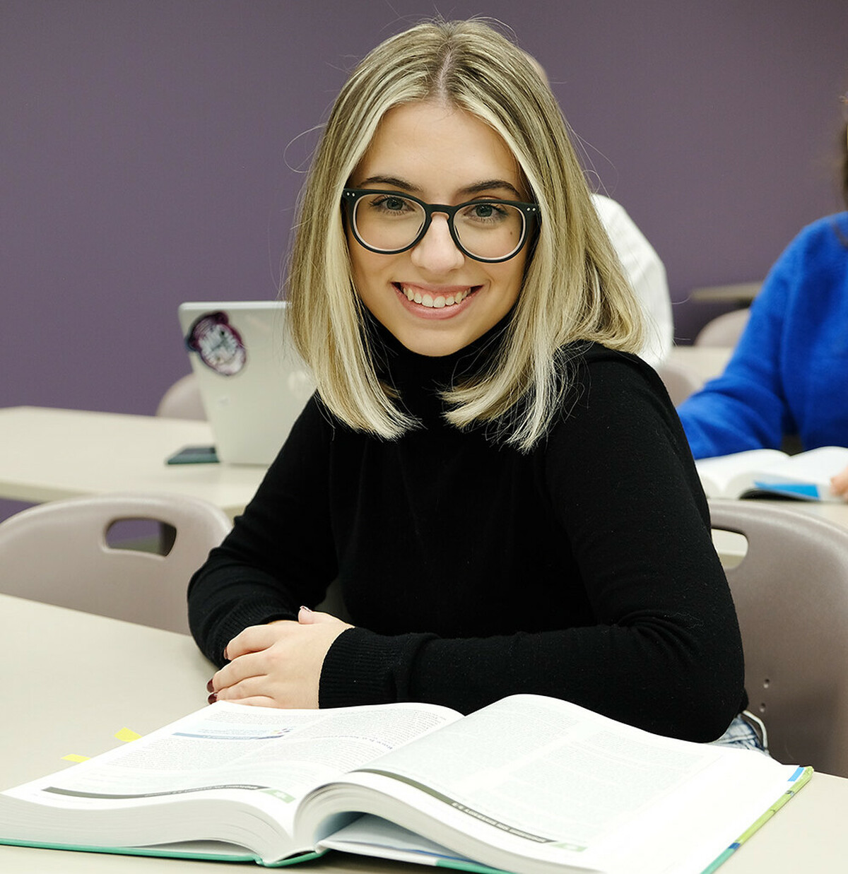 A woman sitting at a desk looks up from her textbook to smile at the camera.
