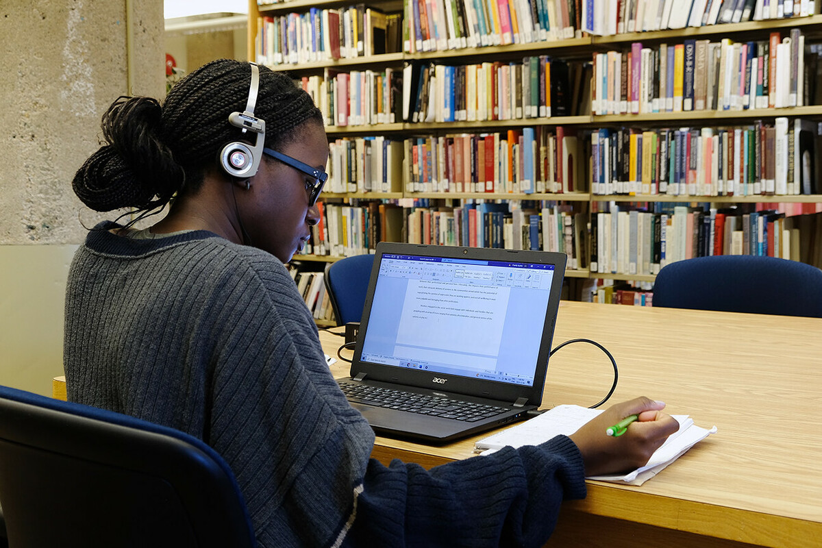 Female student working in a library, writing in a notebook with her laptop open on the desk